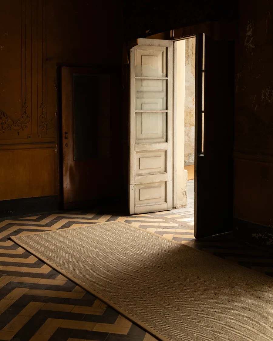 Tapete Lolki 5C con ribete granola sobre piso de duela en espiga, iluminado por una puerta abierta